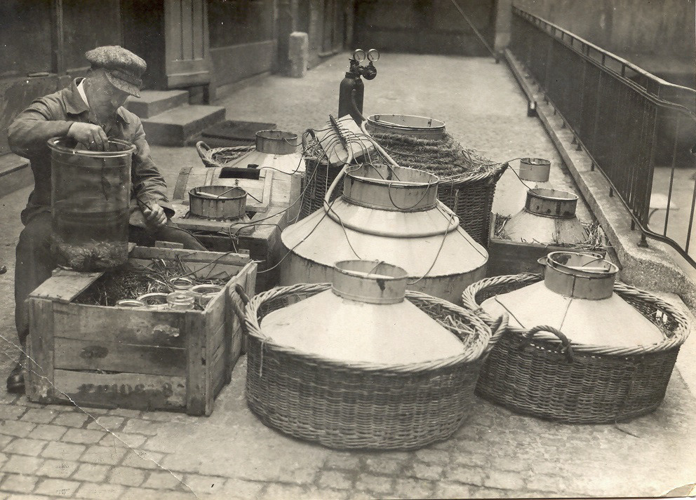Black and white photograph: differently sized baskets, boxes and containers with covers on a paved path. A person sits to the left holding a cylindrical glass container half filled with water.