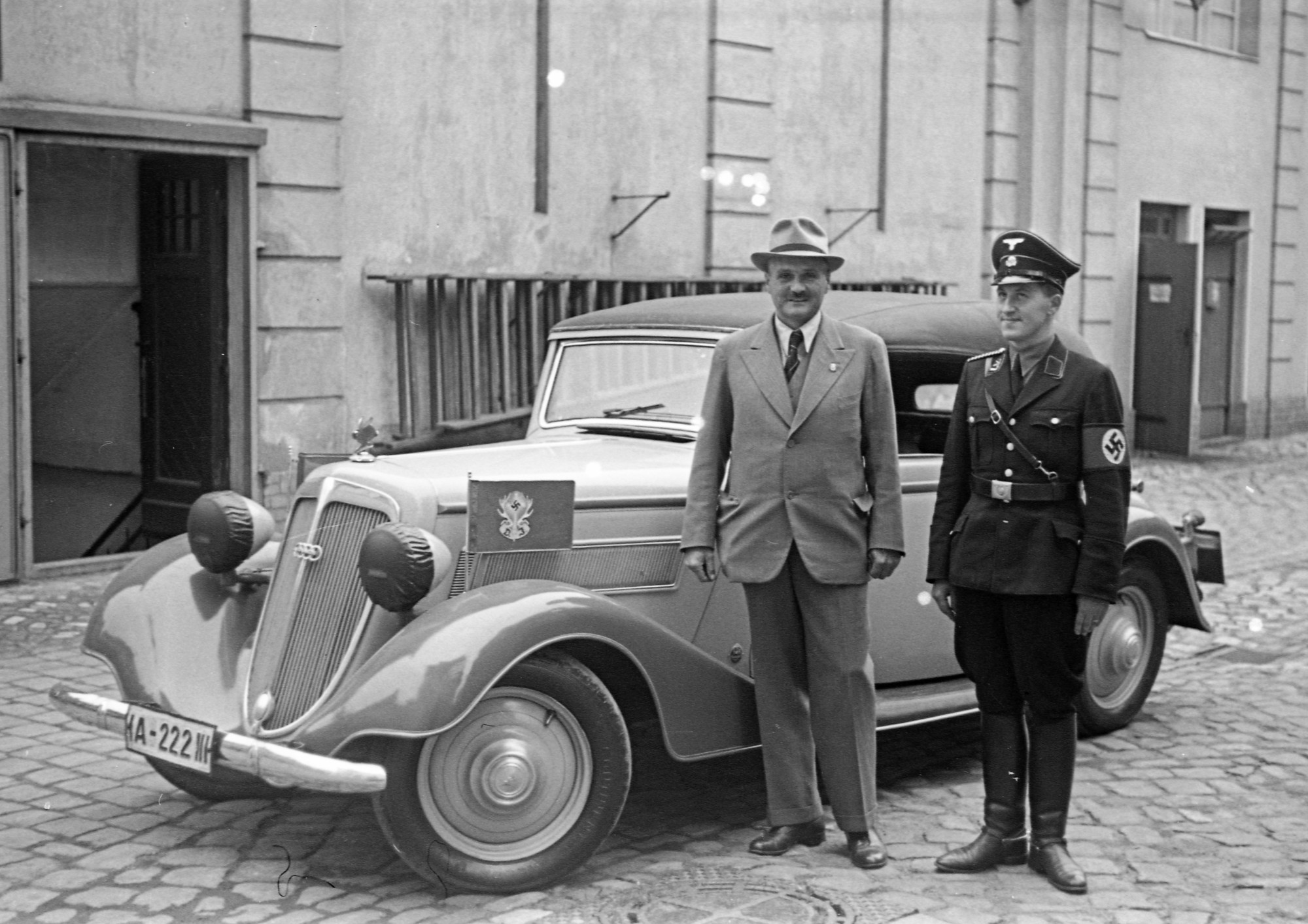 Schwarz-weiß Foto: Mann in Anzug und Hut (li.) sowie Mann in nationalsozialistischer Uniform (re.) stehen lächelnd vor einem geparkten Auto.