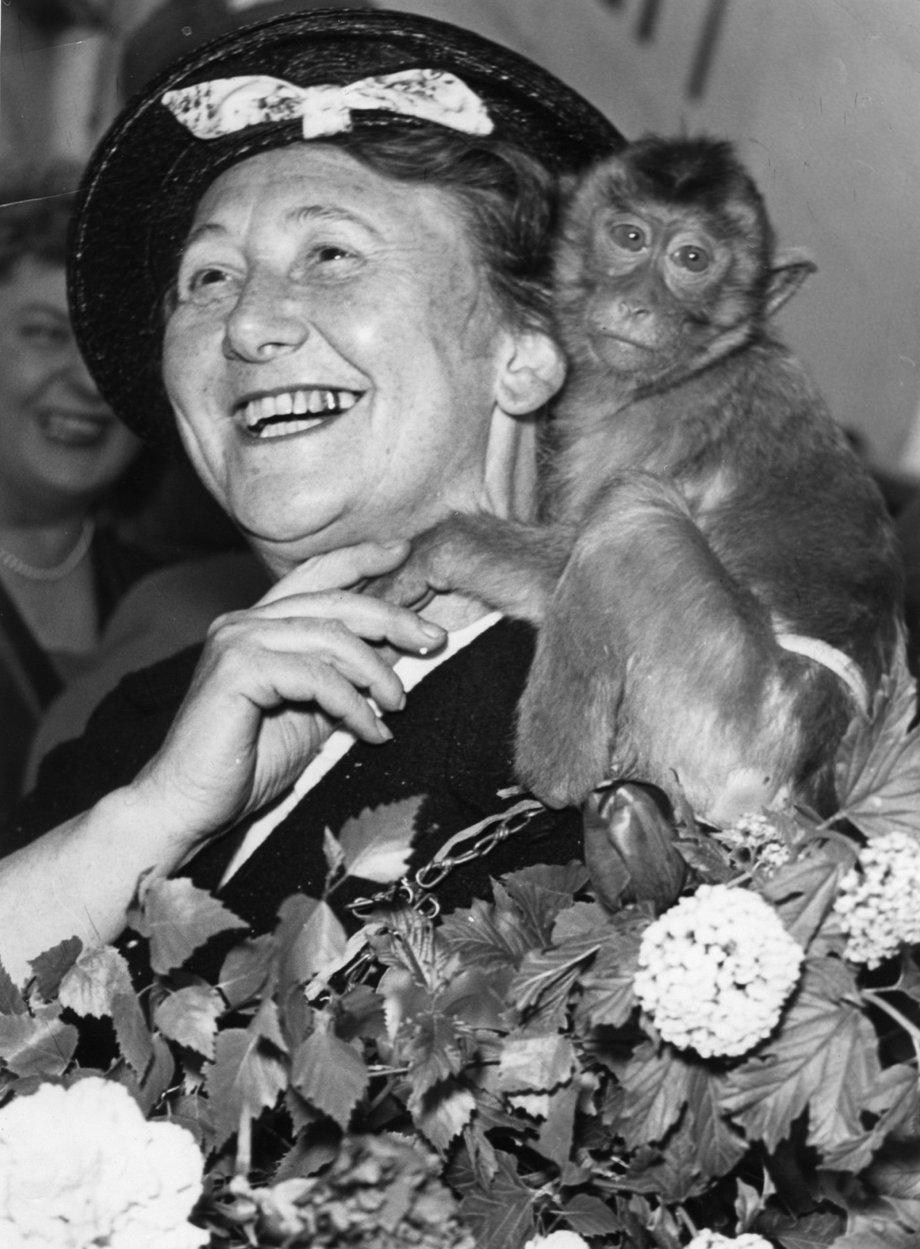 Black and white portrait: Woman in hat, laughing, a monkey on her shoulder. Flower arrangements in the foreground.