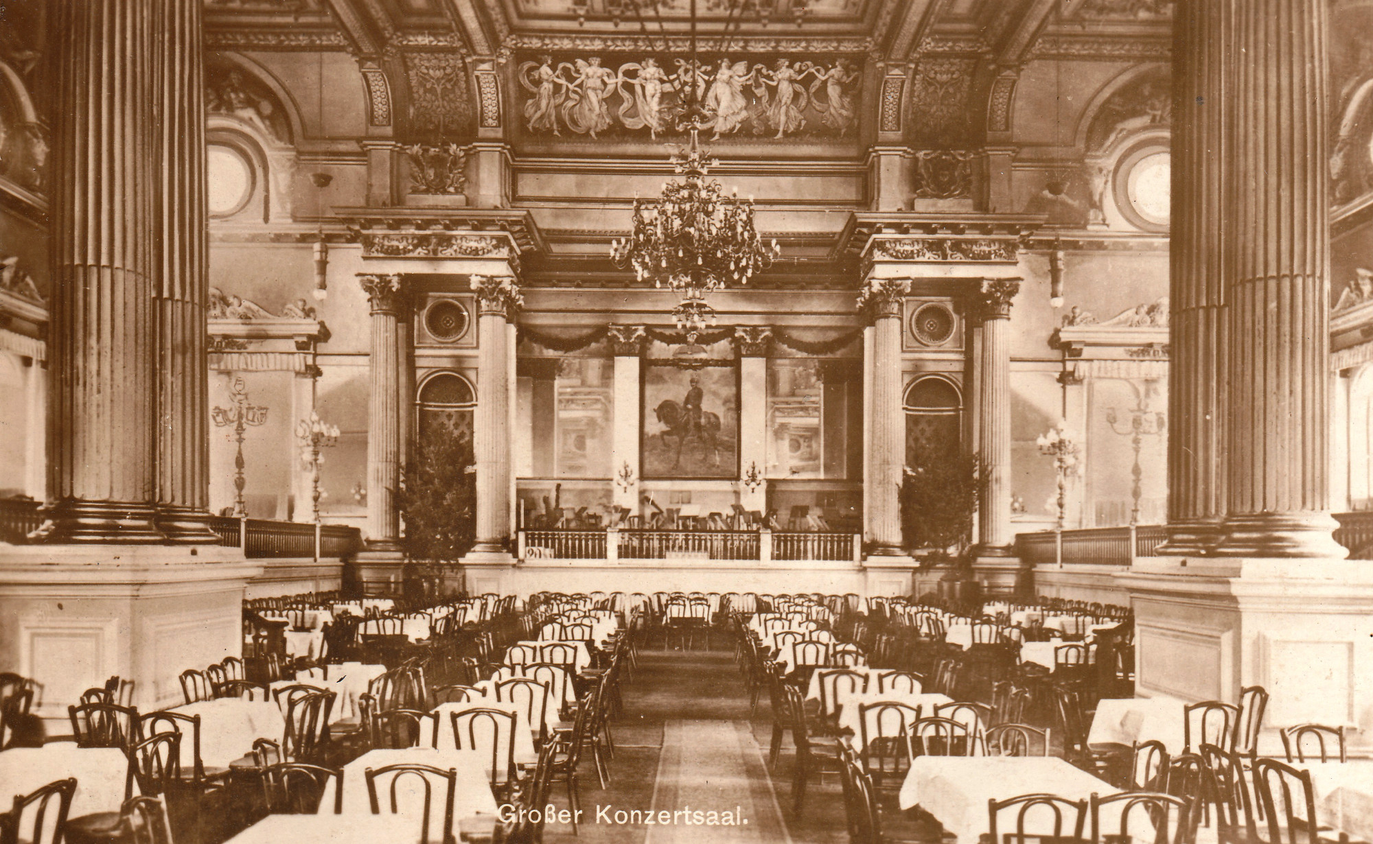 A large, magnificently designed hall with numerous chairs and tables decked in white. The hall reveals various styles, including ornamental ceiling and wall decorations, but also classicist elements such as fluted columns, round windows, and figural patterns. Beneath a giant painting at the end of the hall is a stage with a small balustrade.