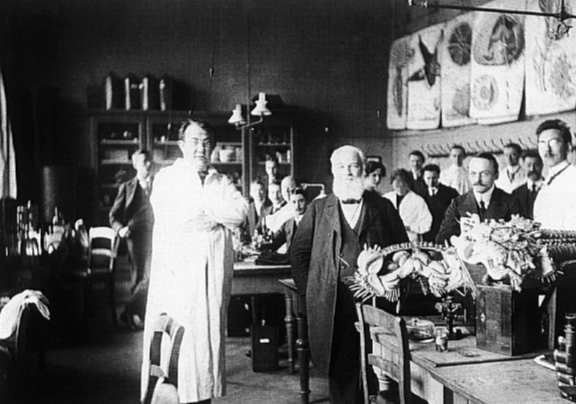 Black and white photo: a classroom with 15 men, many of them with moustaches, and also at least one woman. In the foreground, there is one man on the left in a white lab coat and another on the right with a full white beard wearing a black jacket. At the front, there are objects on a table that are difficult to identify. Five posters of large images hang at the back.