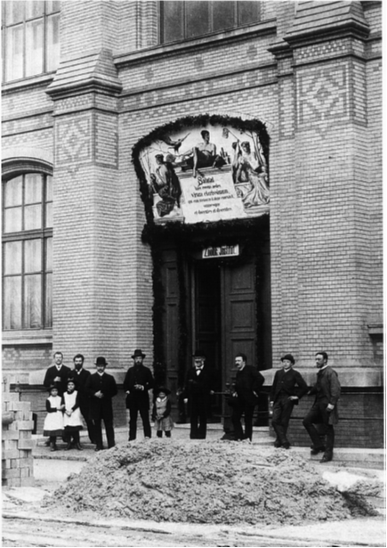 Black and white photo: eight adults and three children stand before the entrance to a brick building, above which a large painting hangs resplendent.