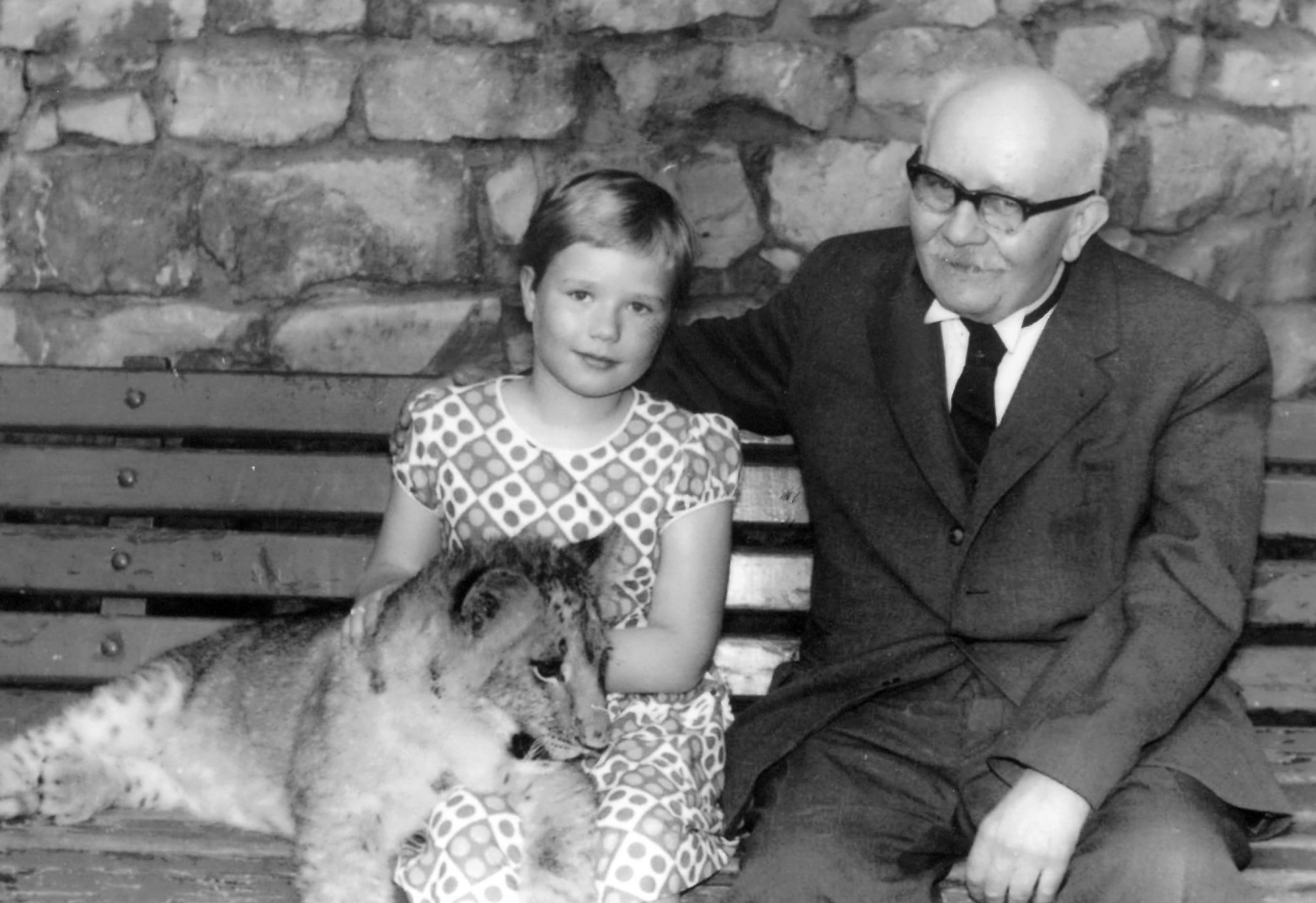 A young girl and an older man sit next to each other on a bench, looking at the camera. There is a lion cub sitting on the girl's lap.