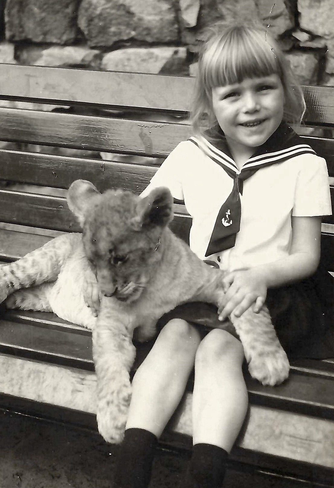 A girl sits smiling on a bench holding a lion cub in her arms.