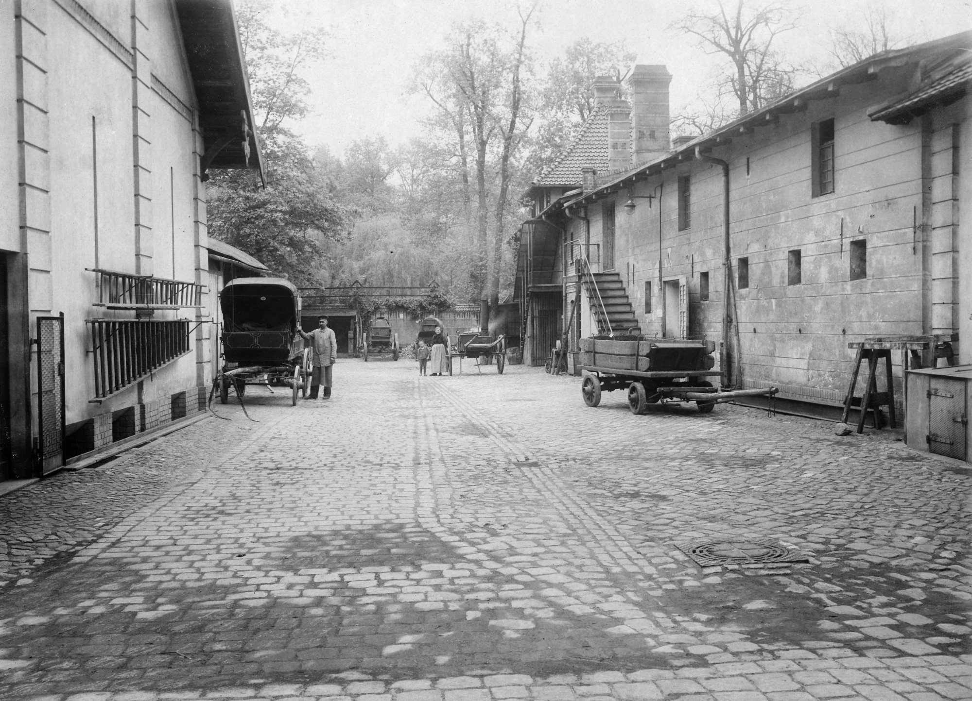 Schwarz-weiß Foto: Gepflasterter, breiter Weg, links und rechts Gebäude, im Hintergrund Bäume. Auf dem Weg steht links ein Mann neben einer Kutsche, rechts und weiter hinten stehen Karren und Arbeitsgeräte.
